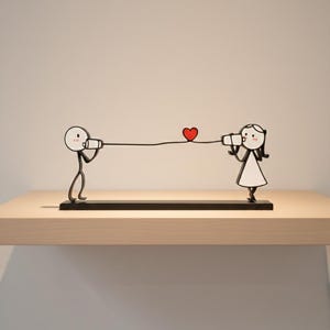 Puede incluir: Escultura de metal negro que representa dos figuras de palo sosteniendo una línea con un corazón rojo en el medio. Las figuras están sobre una base negra, colocada sobre una estantería clara.