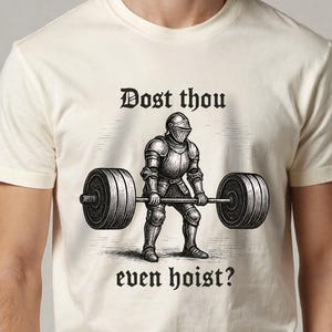 May include: A cream-coloured t-shirt with a black and white illustration of a knight lifting a barbell. The text "Dost thou even hoist?" is above and below the illustration. The shirt is made of a soft material.