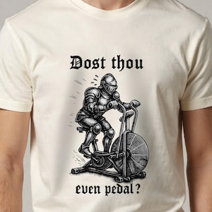 May include: Cream-colored t-shirt featuring a black and white illustration of a knight in full armor riding an exercise bike. The text "Dost thou even pedal?" is above and below the image.