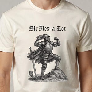 Pu&ograve; includere: T-shirt color crema con un'illustrazione nera di un cavaliere muscoloso in armatura, una spada e la scritta "Sir Flex-a-Lot" in carattere gotico. Il cavaliere &egrave; in piedi su una roccia.