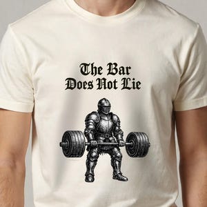 Pode incluir: Camiseta creme com uma ilustração em preto e branco de um cavaleiro levantando uma barra. O texto "The Bar Does Not Lie" está acima do cavaleiro. O desenho está centrado na camiseta.