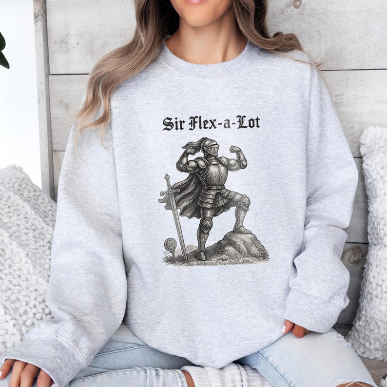 Pu&ograve; includere: Felpa grigio chiaro con la scritta "Sir Flex-a-Lot" sopra un'illustrazione in bianco e nero di un cavaliere che mostra i muscoli. Il cavaliere indossa un'armatura e sta su una roccia con una spada e una mazza.