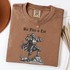 Pu&ograve; includere: Una maglietta marrone con la scritta "Sir Flex-a-Lot" sopra un'illustrazione in bianco e nero di un cavaliere che si flette. La maglietta ha un girocollo e maniche corte. Una collana d'oro &egrave; drappeggiata sulla maglietta.