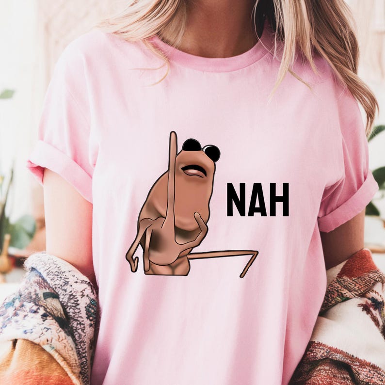 Op de afbeelding: Lichtroze T-shirt met een cartoonillustratie van een bruin wezen met een zwarte zonnebril die een handgebaar maakt. Het woord "NAH" staat in zwarte, vette letters naast het wezen.