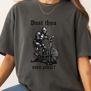 May include: Dark gray t-shirt featuring a black illustration of a knight in full armor riding an exercise bike. The text "Dost thou even pedal?" is above and below the image in a gothic font. The shirt is a relaxed fit.