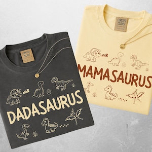 Puede incluir: Dos camisetas, una de color carbón y otra amarilla, con ilustraciones de dinosaurios y texto. La camiseta de color carbón dice "Dadasaurus" y la camiseta amarilla dice "Mamasaurus". Cada camiseta tiene un collar de oro.