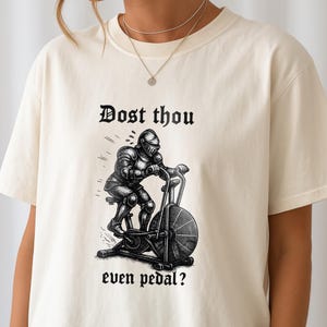 May include: Cream-colored t-shirt featuring a black and white illustration of a knight on a stationary bike. The text "Dost thou even pedal?" is printed above and below the image in a gothic font. The shirt is made of a soft material.