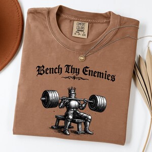 May include: A brown t-shirt with the text "Bench Thy Enemies" above an illustration of a knight lifting weights. The shirt has a crew neck and a small tag on the upper right. A silver necklace with a round pendant is draped across the shirt.