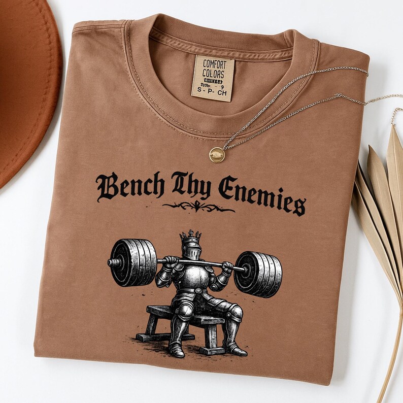 May include: A brown t-shirt with the text "Bench Thy Enemies" above an illustration of a knight lifting weights. The shirt has a crew neck and a small tag on the upper right. A silver necklace with a round pendant is draped across the shirt.