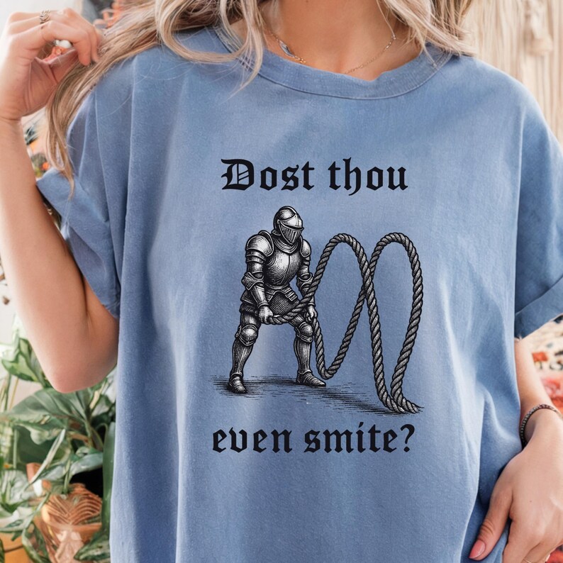 Puede incluir: Camiseta azul claro con un gr&aacute;fico de caballero medieval y el texto "Dost thou even smite?" en fuente g&oacute;tica. La ilustraci&oacute;n muestra un caballero con armadura sosteniendo una cuerda. La camiseta tiene mangas cortas.