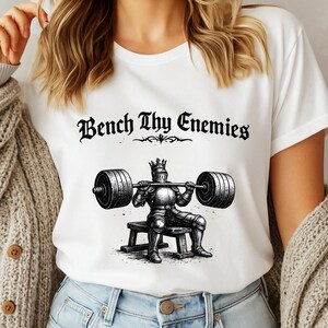 May include: White t-shirt featuring a black and white illustration of a knight lifting weights. The knight is wearing armor and a crown. The text "Bench Thy Enemies" is above the illustration.