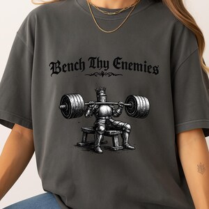 May include: Dark grey t-shirt with the text "Bench Thy Enemies" above an illustration of a knight lifting a barbell. The knight is wearing armor and a crown, seated on a bench. The design is in black and white.