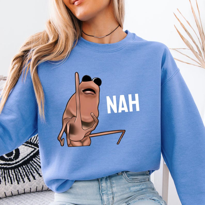 Op de afbeelding: Lichtblauw sweatshirt met een cartoonachtig bruin wezen met een zonnebril en het woord "NAH" in witte letters. Het wezen steekt zijn middelvinger op. Het sweatshirt is effen lichtblauw.