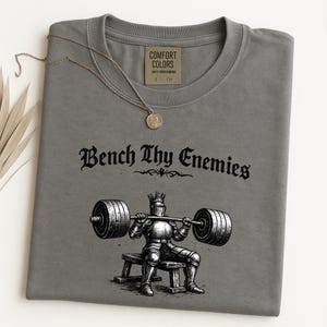 May include: A gray Comfort Colors t-shirt with the text "Bench Thy Enemies" above an illustration of a knight lifting weights. A gold-colored necklace with a pendant is draped over the shirt.