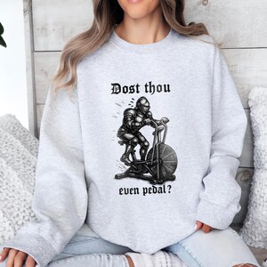 May include: Light gray sweatshirt with the text "Dost thou even pedal?" above a black and white illustration of a knight on an exercise bike. The sweatshirt is a crew neck style.