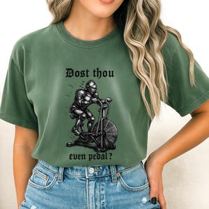 May include: Green t-shirt with the text "Dost thou even pedal?" above a graphic of a knight in armor riding an exercise bike. The shirt is a solid green color and has a crew neck.