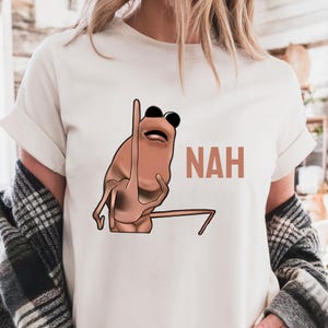Op de afbeelding: Cr&egrave;mekleurig T-shirt met een cartoonachtig bruin wezen met een zwarte zonnebril en een opgeheven arm. Het woord "NAH" is in bijpassende bruine kleur aan de rechterkant van het shirt gedrukt. Het shirt heeft korte mouwen.