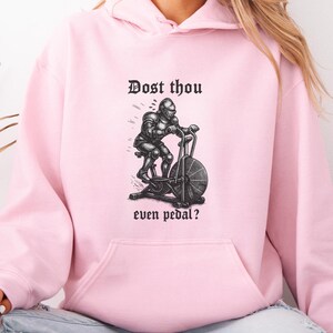 May include: A light pink hoodie featuring a black and white illustration of a knight on an exercise bike. The text "Dost thou even pedal?" is printed above and below the illustration in a gothic font.
