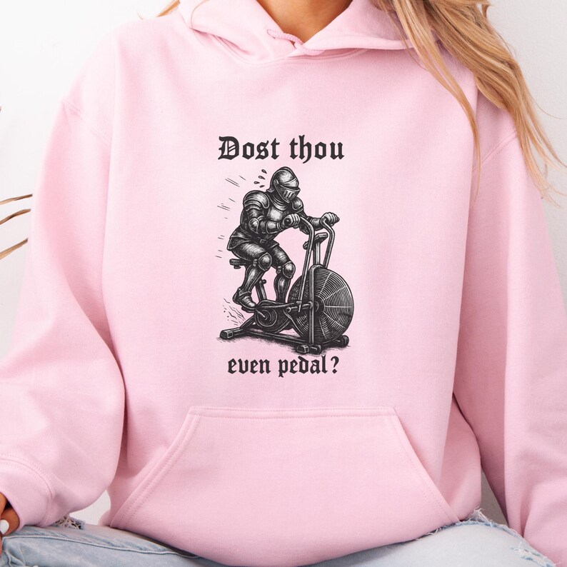 May include: A light pink hoodie featuring a black and white illustration of a knight on an exercise bike. The text "Dost thou even pedal?" is printed above and below the illustration in a gothic font.