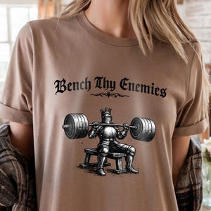 May include: A tan t-shirt featuring the phrase "Bench Thy Enemies" in a gothic font above an illustration of a knight bench pressing a barbell. The knight is wearing armor and a crown. The barbell has weight plates on each end.