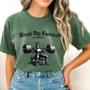 May include: Green t-shirt with the text "Bench Thy Enemies" above a graphic of a knight lifting weights. The knight is wearing armor and a crown. The shirt is a solid green color.