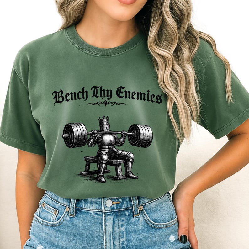 May include: Green t-shirt with the text "Bench Thy Enemies" above a graphic of a knight lifting weights. The knight is wearing armor and a crown. The shirt is a solid green color.