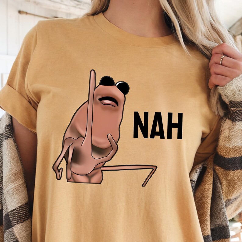 Op de afbeelding: Mosterdgeel T-shirt met een stripfiguur met zwarte zonnebril en het woord "NAH" in zwarte blokletters. Het personage is lichtbruin en maakt een afwijzend gebaar.
