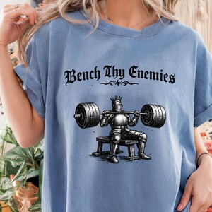 May include: Light blue t-shirt with the text "Bench Thy Enemies" in a gothic font. The graphic depicts a knight in armor bench pressing a barbell. The shirt has short sleeves and a relaxed fit.