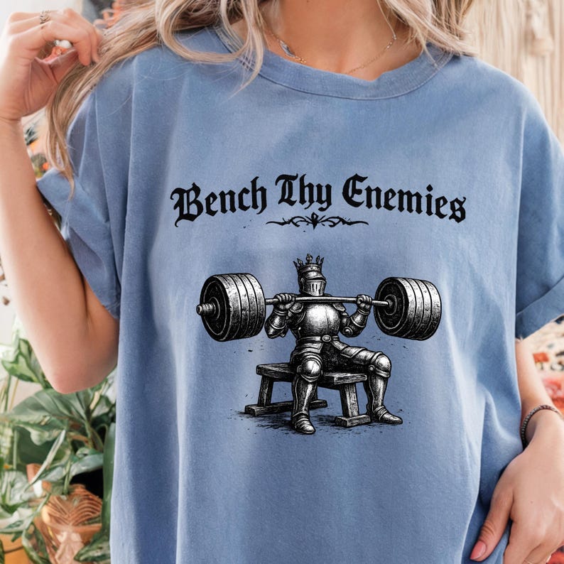 May include: Light blue t-shirt with the text "Bench Thy Enemies" in a gothic font. The graphic depicts a knight in armor bench pressing a barbell. The shirt has short sleeves and a relaxed fit.