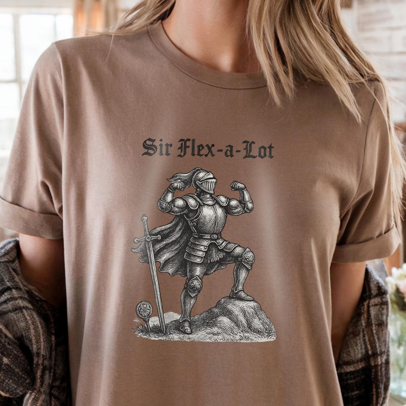 Pu&ograve; includere: T-shirt color cachi con la scritta "Sir Flex-a-Lot" sopra un'illustrazione in bianco e nero di un cavaliere muscoloso. Il cavaliere indossa un'armatura e sta in piedi su una roccia con una spada al suo fianco.