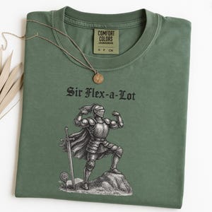 Pu&ograve; includere: T-shirt verde oliva con la scritta "Sir Flex-a-Lot" e una grafica di un cavaliere che mostra i muscoli. Una collana color oro con un ciondolo rotondo &egrave; appoggiata sulla maglietta. La maglietta ha un'etichetta Comfort Colors.
