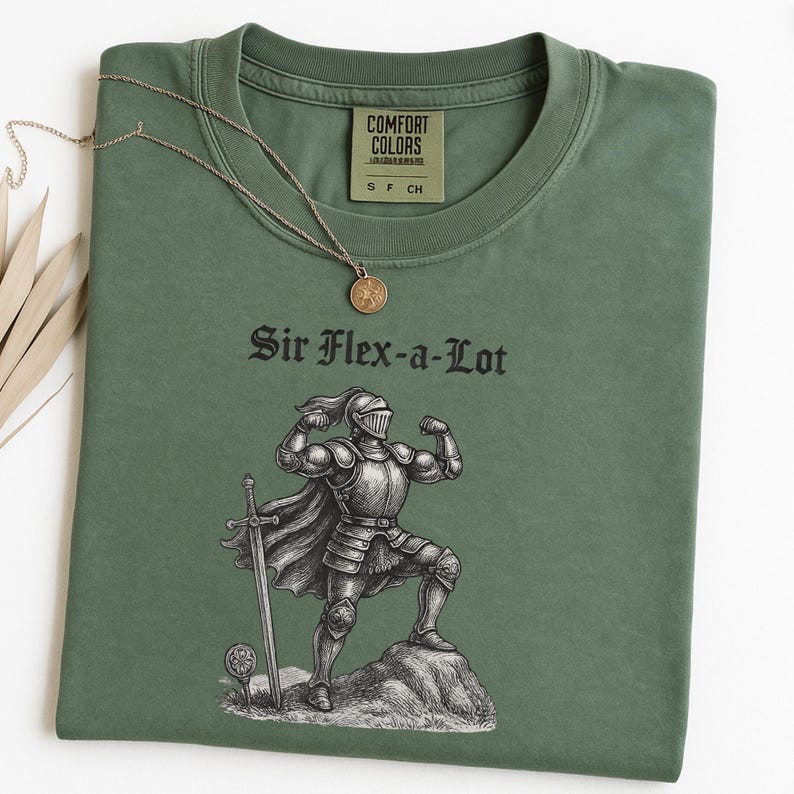 Pu&ograve; includere: T-shirt verde oliva con la scritta "Sir Flex-a-Lot" e una grafica di un cavaliere che mostra i muscoli. Una collana color oro con un ciondolo rotondo &egrave; appoggiata sulla maglietta. La maglietta ha un'etichetta Comfort Colors.