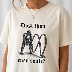 Puede incluir: Camiseta color crema con un dise&ntilde;o gr&aacute;fico en blanco y negro de un caballero medieval sosteniendo una cuerda y el texto "Dost thou even smite?". El dise&ntilde;o es de estilo medieval.