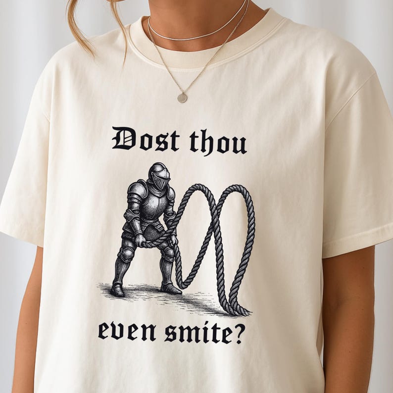 Puede incluir: Camiseta color crema con un dise&ntilde;o gr&aacute;fico en blanco y negro de un caballero medieval sosteniendo una cuerda y el texto "Dost thou even smite?". El dise&ntilde;o es de estilo medieval.