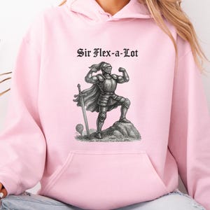 Pu&ograve; includere: Felpa con cappuccio rosa chiaro con tasca frontale e grafica di un cavaliere muscoloso in armatura, una spada e la scritta "Sir Flex-a-Lot" in nero.
