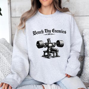 May include: A light gray sweatshirt with the text "Bench Thy Enemies" above a black and white illustration of a knight lifting weights. The knight is wearing a crown and sitting on a bench.