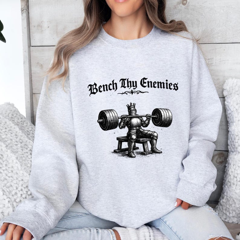May include: A light gray sweatshirt with the text "Bench Thy Enemies" above a black and white illustration of a knight lifting weights. The knight is wearing a crown and sitting on a bench.