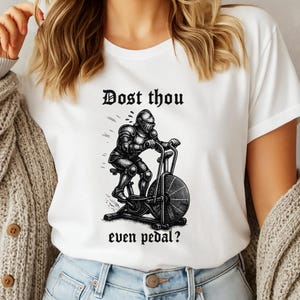 May include: White t-shirt with a black graphic of a knight on an exercise bike. The text "Dost thou even pedal?" is above and below the illustration. The shirt is made of a soft material.