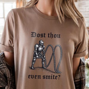 Puede incluir: Camiseta color beige con un dise&ntilde;o en blanco y negro de un caballero con armadura sosteniendo una cuerda. El texto "Dost thou even smite?" est&aacute; impreso arriba y abajo de la imagen en fuente g&oacute;tica.