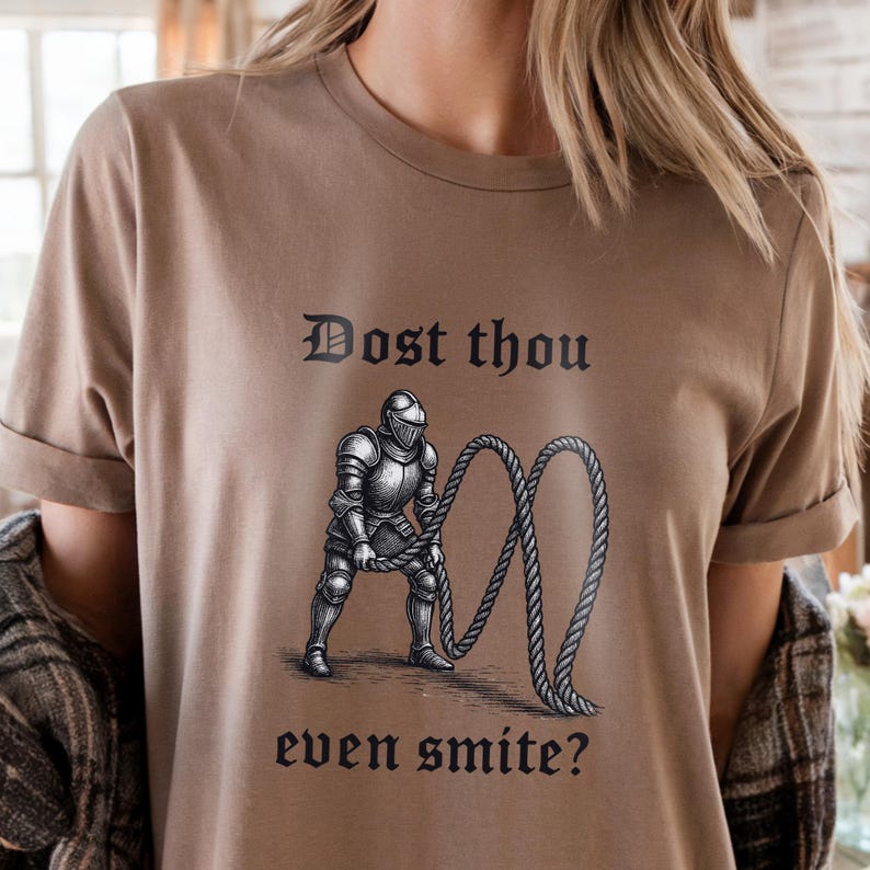 Puede incluir: Camiseta color beige con un dise&ntilde;o en blanco y negro de un caballero con armadura sosteniendo una cuerda. El texto "Dost thou even smite?" est&aacute; impreso arriba y abajo de la imagen en fuente g&oacute;tica.