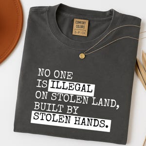May include: Dark grey t-shirt with the text "NO ONE IS ILLEGAL ON STOLEN LAND, BUILT BY STOLEN HANDS." A gold necklace and a brown hat are visible. The shirt has a Comfort Colors tag.