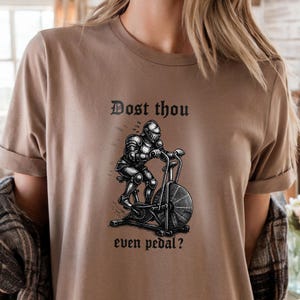 May include: A tan t-shirt with the text "Dost thou even pedal?" above a black and white illustration of a knight on an exercise bike. The shirt has rolled-up sleeves.