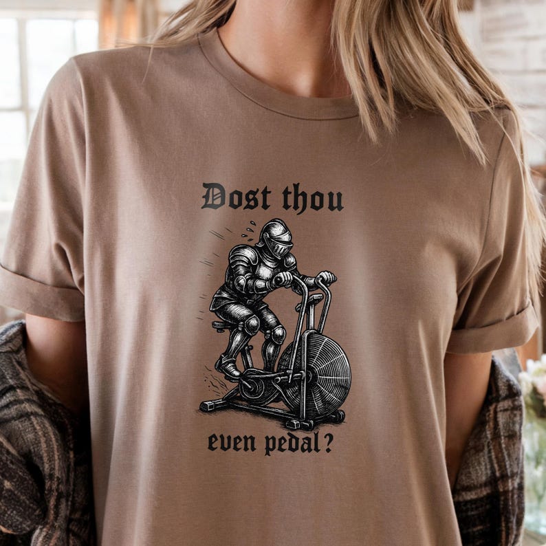 May include: A tan t-shirt with the text "Dost thou even pedal?" above a black and white illustration of a knight on an exercise bike. The shirt has rolled-up sleeves.
