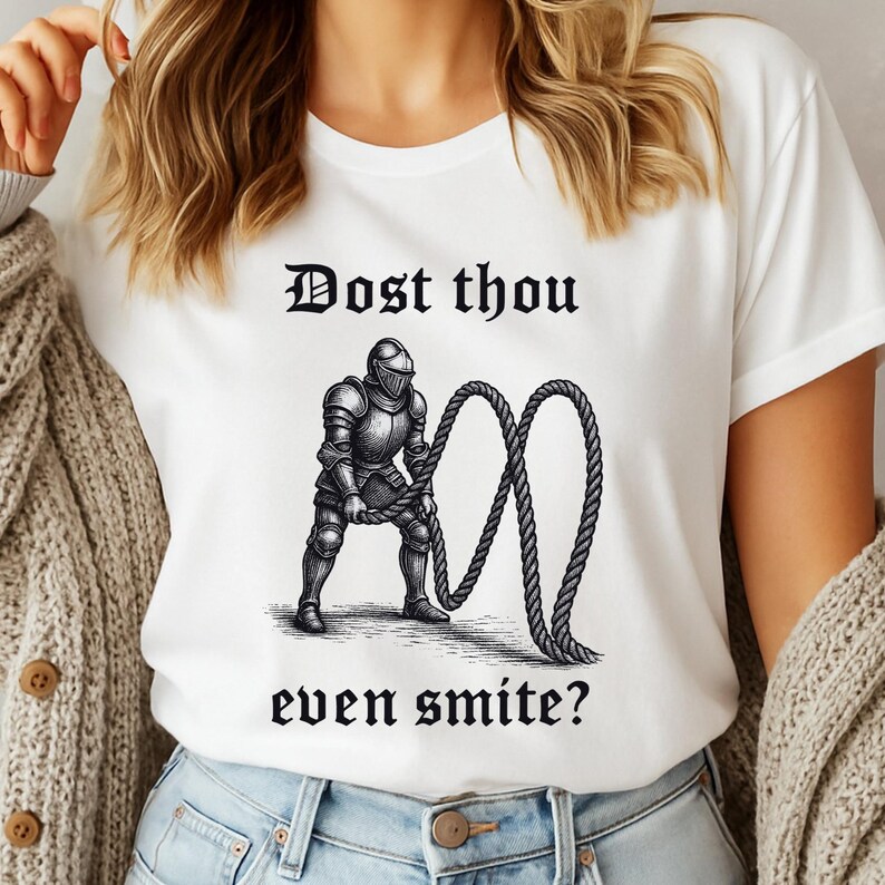 Puede incluir: Camiseta blanca con una ilustraci&oacute;n en blanco y negro de un caballero sosteniendo una cuerda, con el texto "Dost thou even smite?" en fuente g&oacute;tica negra. El dise&ntilde;o est&aacute; centrado en la camiseta.