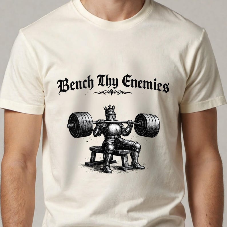 May include: Cream-colored t-shirt featuring a black illustration of a knight lifting weights. The knight is wearing a crown and armor, seated on a bench. The text "Bench Thy Enemies" is above the illustration in a gothic font.