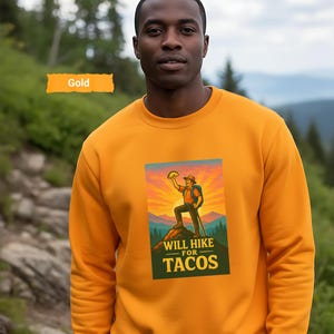 May include: Gold-colored sweatshirt featuring a graphic that reads "WILL HIKE FOR TACOS." The graphic depicts a hiker holding a taco with a mountain backdrop. The sweatshirt is worn by a person standing on a trail.