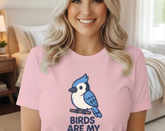 May include: Pink t-shirt featuring a cartoon blue jay perched on a branch, with the text "BIRDS ARE MY THING" in navy blue. The shirt is a crew neck style.