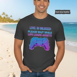 May include: Dark grey heather t-shirt featuring a neon blue and purple video game controller graphic. The shirt has the text "LEVEL 50 UNLOCKED: PLEASE WAIT WHILE LIFE LOADS ASSETS". The shirt is worn by a person on a beach.