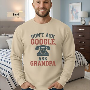 May include: Sand-colored sweatshirt with the text "DON'T ASK GOOGLE, ASK GRANDPA" in red and blue. The design includes a vintage telephone graphic. The sweatshirt is a casual style.