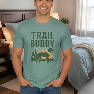 May include: Light green t-shirt with the text "TRAIL BUDDY" above a graphic of a dog, mountains, and a sunburst. The shirt is worn by a person in blue jeans.